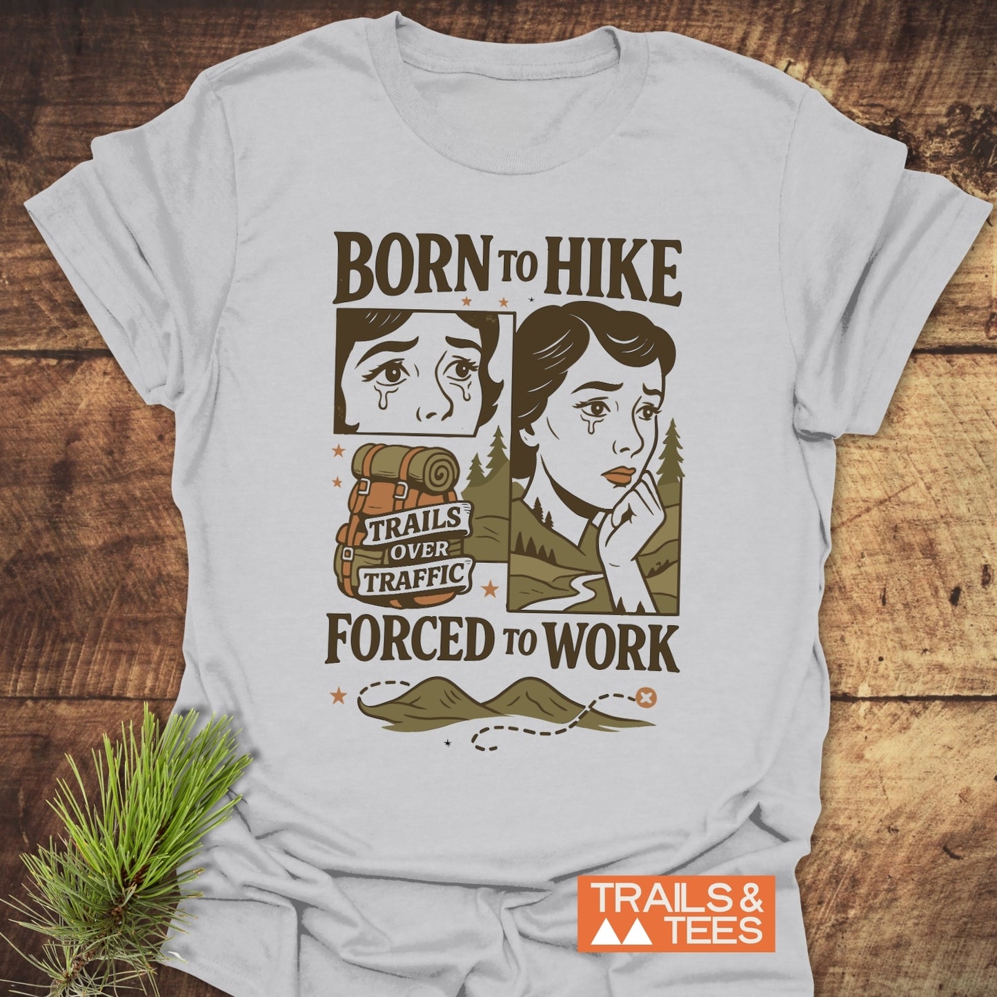 Light-gray Forced To Work Funny T-Shirt with hiking-themed comic print, featuring a hiker, rucksack, and TRAILS & TEES logo, displayed on a wooden surface.