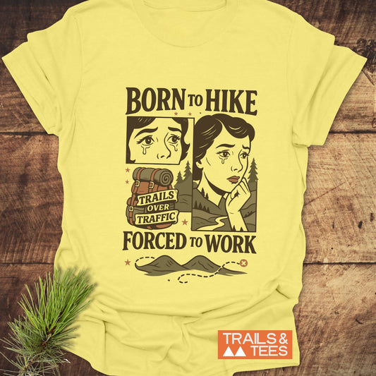 Forced To Work Funny T-Shirt with comic-style “BORN TO HIKE” design, features teary-eyed woman, hiking pack, mountain path, and “TRAILS & TEES” logo.