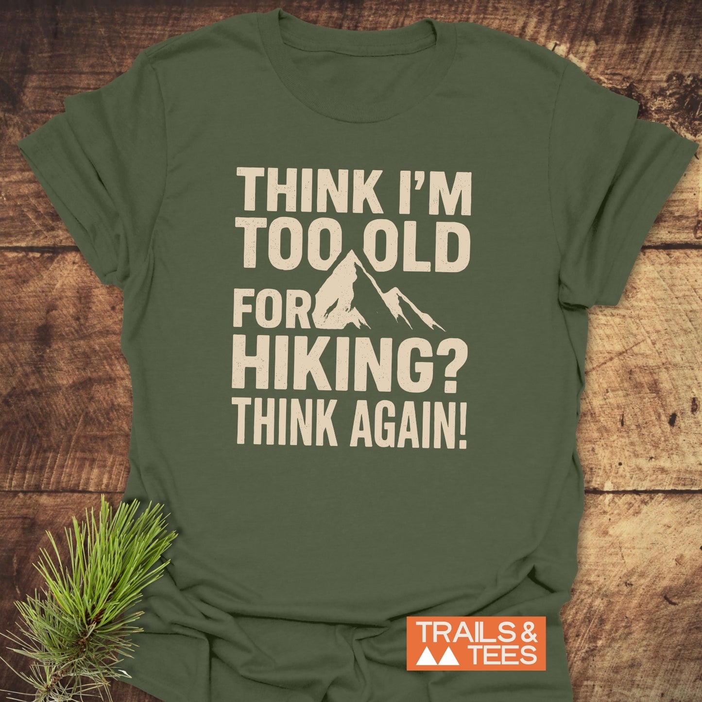 Olive-green Think Again Funny T-Shirt with cream text and mountain silhouette, laid on wood. Made from ring-spun cotton, offers durability and comfort for outdoor enthusiasts.