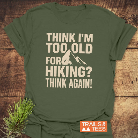 Olive-green Think Again Funny T-Shirt with cream text and mountain silhouette, laid on wood. Made from ring-spun cotton, offers durability and comfort for outdoor enthusiasts.