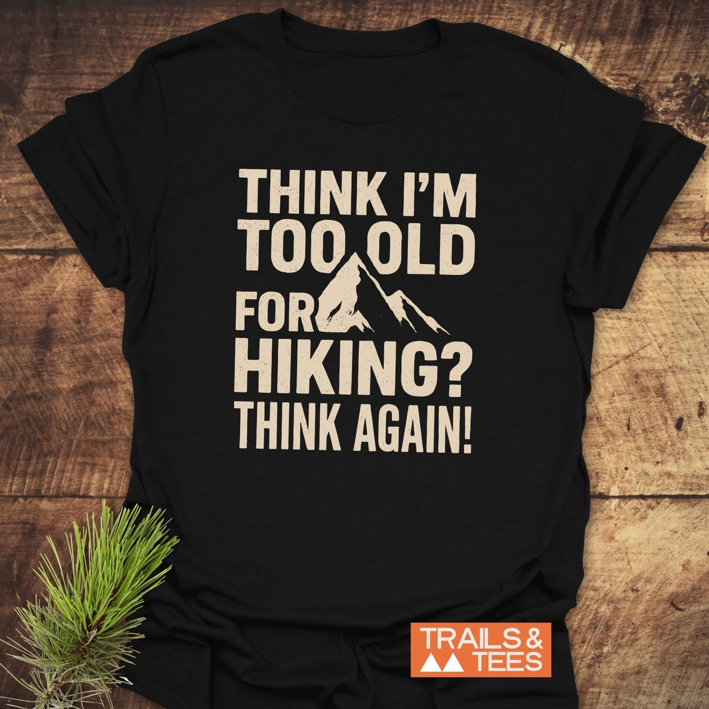 Black Think Again Funny T-Shirt with mountain graphic, slogan about hiking, laid on wood; includes TRAILS & TEES logo. Crafted for outdoor enthusiasts.