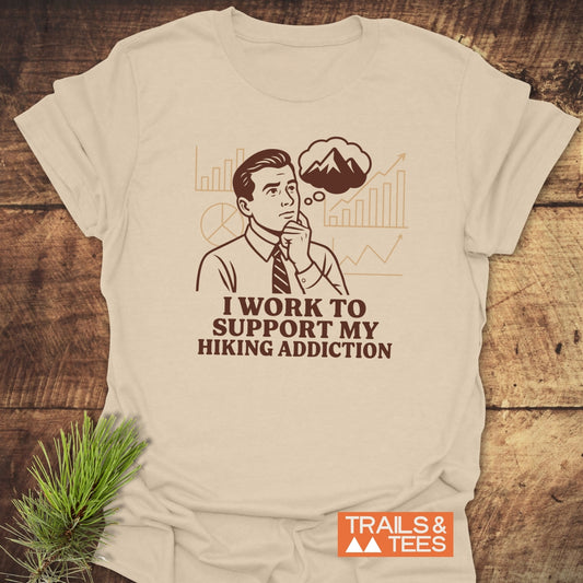 Hiking Addiction Funny T-Shirt featuring a retro business man with mountain thought bubble, slogan, and durable design, styled with pine sprig on wooden surface.