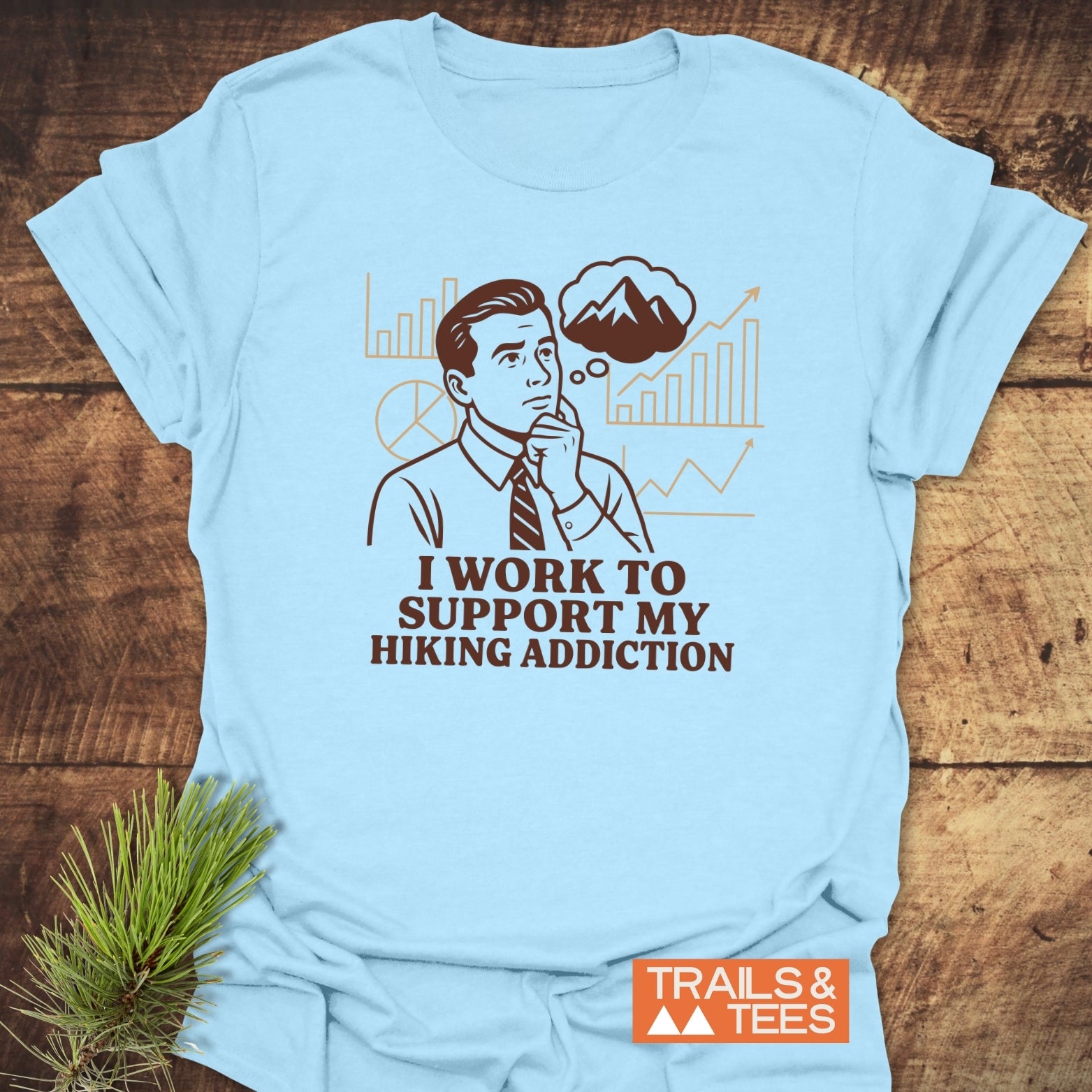 Light-blue Hiking Addiction Funny T-Shirt with retro illustration, text I WORK TO SUPPORT MY HIKING ADDICTION, and TRAILS & TEES logo, laid on wooden surface.