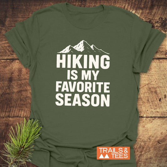 Olive-green t-shirt with “HIKING IS MY FAVORITE SEASON” slogan, featuring durable print, laid on wood. Includes “TRAILS & TEES” logo and pine branch accent.