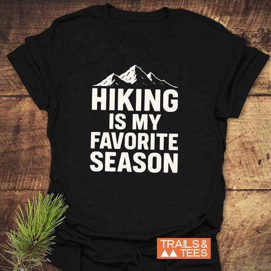 Hiking Is My Favorite Season T-Shirt featuring mountain graphic and Trails & Tees logo, laid on wood surface with pine needles, emphasizes outdoor adventure theme.