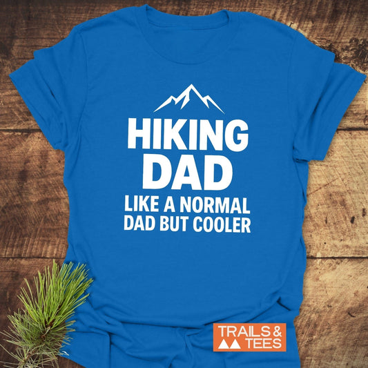 Royal-blue Hiking Dad T-shirt with mountain design, white text, and orange TRAILS & TEES logo, laid on rustic wood with pine sprig nearby.