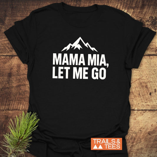 Let Me Go Funny T-Shirt: Black tee with mountain silhouette and slogan, laid on wood, evokes outdoor adventure with Trails & Tees logo and pine branch accent.