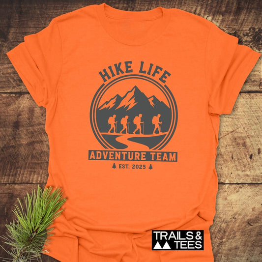 Adventure Team T-Shirt featuring HIKE LIFE arc, mountain range, backpackers, and ADVENTURE TEAM banner, laid flat on wood with pine sprig, showcasing outdoor spirit.