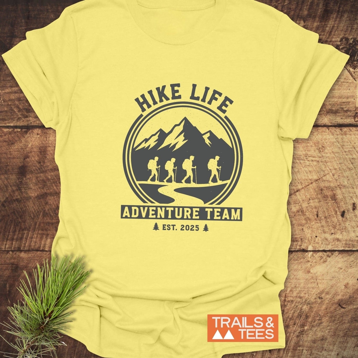 Adventure Team T-Shirt featuring a “HIKE LIFE” badge design with hikers and mountains, emphasizing comfort, durability, and outdoor spirit. Includes Trails & Tees logo.