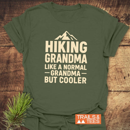 Hiking Grandma T-Shirt featuring a mountain icon and slogan, displayed on a rustic backdrop with pine branch, showcasing outdoor theme and comfort design.