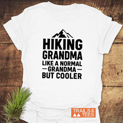Hiking Grandma T-Shirt featuring a mountain silhouette with bold text, displayed on wood with a pine sprig and Trails & Tees logo.