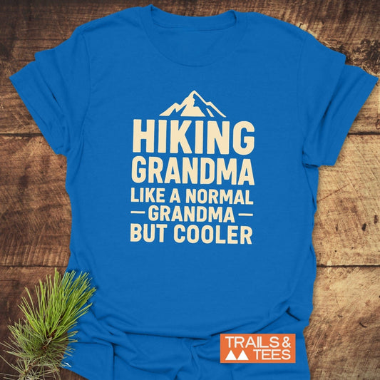 Hiking Grandma T-Shirt with mountain graphic, laid on wood. Cream print: “Hiking Grandma, Like a Normal Grandma, But Cooler.” Includes Trails & Tees logo.