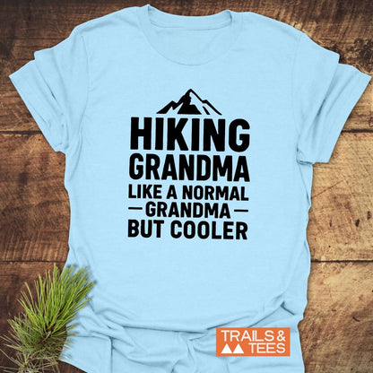 Light blue Hiking Grandma T-Shirt with a mountain icon, bold text, and Trails & Tees logo, on a wooden surface with a pine sprig.