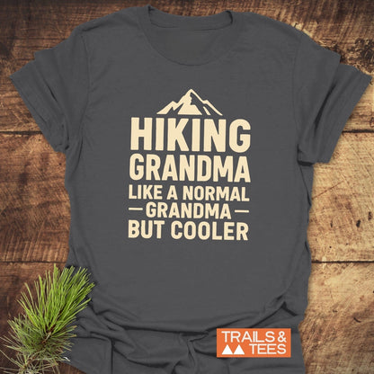 Dark gray Hiking Grandma T-Shirt with mountain silhouette and slogan, styled on wood with pine sprig, featuring TRAILS & TEES logo, emphasizing comfort and adventure.