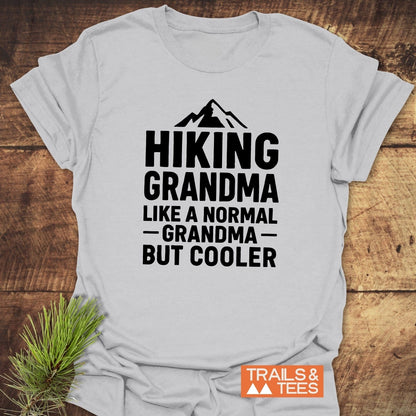 Light-gray Hiking Grandma T-Shirt with mountain silhouette and bold text, laid on wood. Pine sprig and TRAILS & TEES logo beside it.