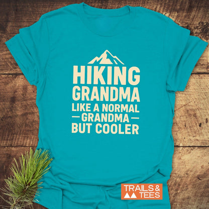 Turquoise T-shirt with “HIKING GRANDMA” print, featuring a mountain silhouette. Includes Trails & Tees logo and pine sprig accent for rustic appeal.