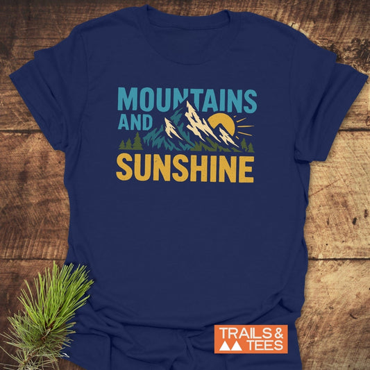 Mountains And Sunshine T-Shirt featuring a stylized mountain, sun, and pine design, emphasizing outdoor adventure. Includes TRAILS & TEES logo and pine sprig accent.