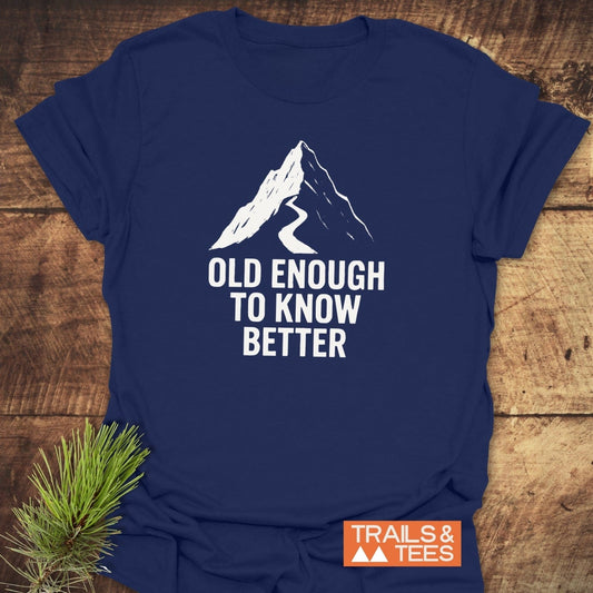 Old Enough Funny T-Shirt featuring a mountain design and slogan on a navy-blue tee, laid flat with a pine sprig, showcasing comfort and outdoor spirit.
