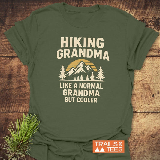 Hiking Grandma Sunrise T-Shirt featuring a sunrise behind a mountain with pine trees, text: HIKING GRANDMA, LIKE A NORMAL GRANDMA BUT COOLER, on soft cotton.