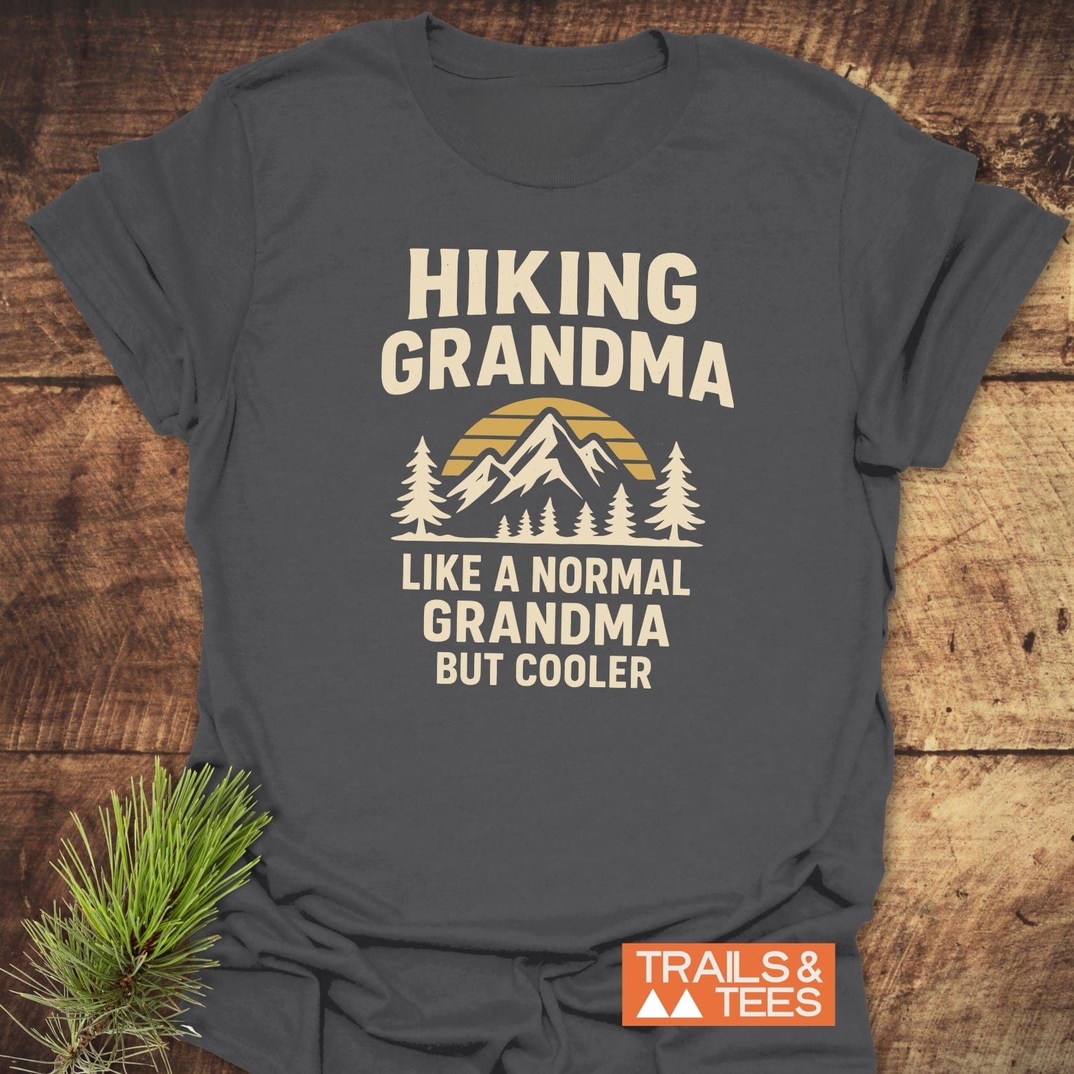 Hiking Grandma Sunrise T-Shirt: dark-gray tee with “HIKING GRANDMA” and mountain graphic. Cream print. Durable, soft cotton, ideal for outdoor enthusiasts.