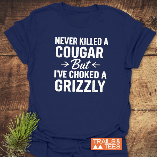 Never Killed A Cougar T-Shirt on wooden surface, featuring bold slogan, Trails & Tees logo, crafted from soft cotton for outdoor adventures.