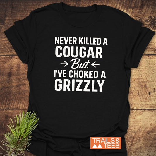 Never Killed A Cougar T-Shirt on wood, slogan in white. Features soft, lightweight cotton, taped shoulders, durable print. Includes Trails & Tees logo.