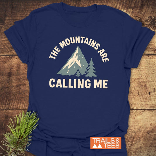 Navy blue Mountains Calling Me T-Shirt with snowy mountain design, pine trees, slogan, and TRAILS & TEES logo, placed on wood with pine sprig.