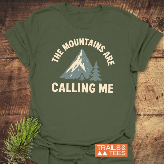 Mountains Calling Me T-Shirt featuring a mountain and pine-tree graphic, laid on wood. Includes TRAILS & TEES logo, with a pine sprig detail.