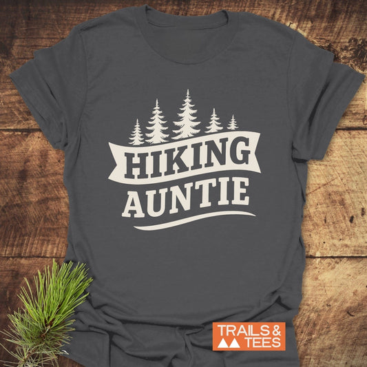 Hiking Auntie T-Shirt on wooden surface, featuring cream pine tree graphic. Soft ring-spun cotton, durable print, taped shoulders, pre-shrunk for reliable fit.