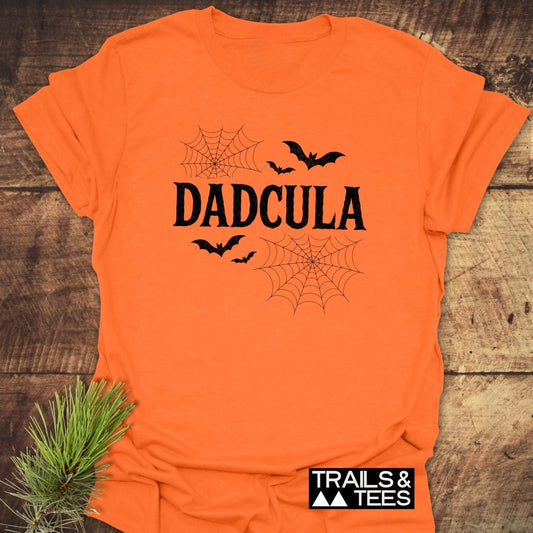 Dadcula Halloween T-Shirt featuring 'DADCULA' text with bats and spider-webs, lying flat with evergreen sprig; soft, durable cotton ideal for outdoor adventures.