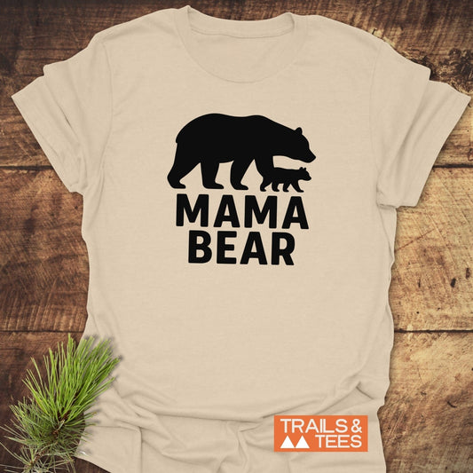Mama Bear T-Shirt with black bear silhouette, “MAMA BEAR” text, and Trails & Tees logo, laid on rustic wood with pine sprig, showcasing outdoor-inspired design.