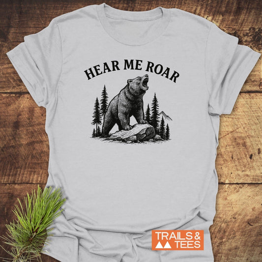 Hear Me Roar T-Shirt featuring a black graphic of a roaring bear on a rock with trees and mountains, laid on wood, beside a pine sprig.