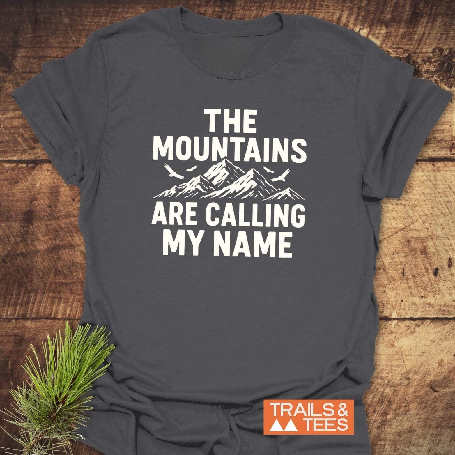 Calling My Name T-Shirt featuring a mountain graphic and birds, with “TRAILS & TEES” logo, laid on wood with a pine sprig.