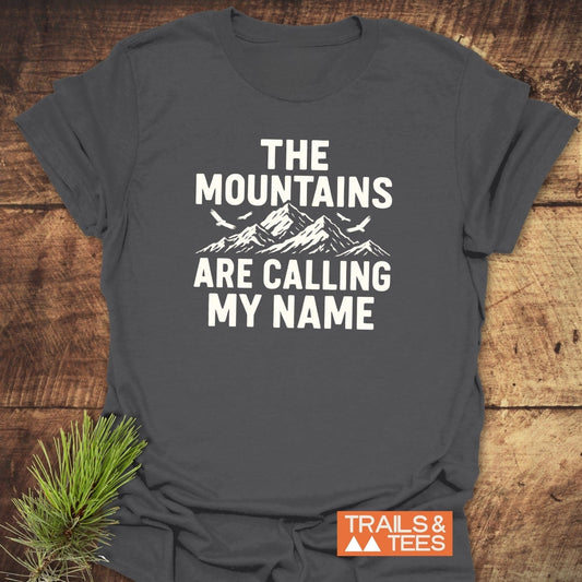 Calling My Name T-Shirt featuring a mountain graphic and birds, with “TRAILS & TEES” logo, laid on wood with a pine sprig.