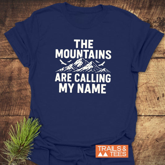 Calling My Name T-Shirt features a mountain graphic and text, emphasizing outdoor adventures. Includes a “TRAILS & TEES” logo, styled with a pine branch.