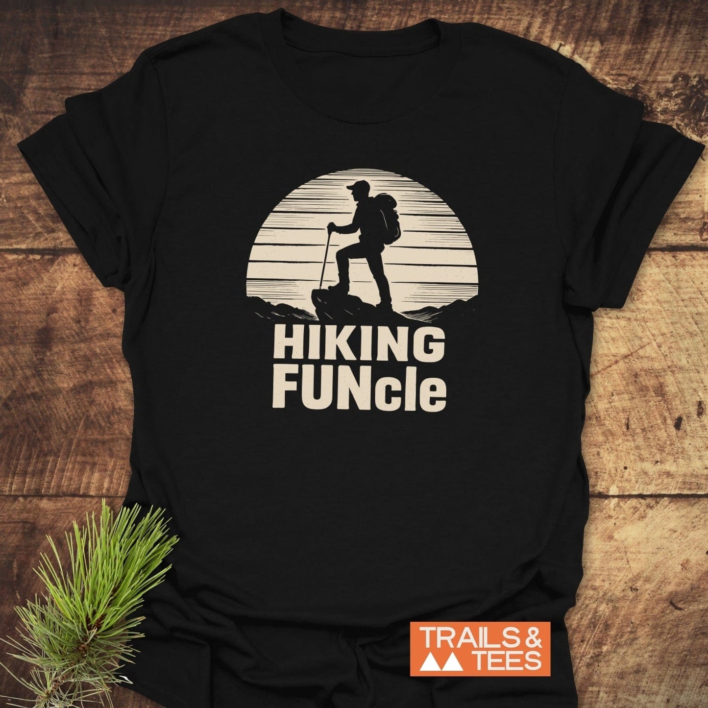 Hiking FUNcle T-Shirt with a hiker graphic and text, laid flat on wood. Includes brand logo and pine sprig accent.
