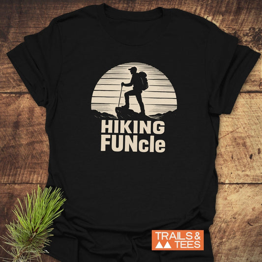 Hiking FUNcle T-Shirt with a hiker graphic and text, laid flat on wood. Includes brand logo and pine sprig accent.