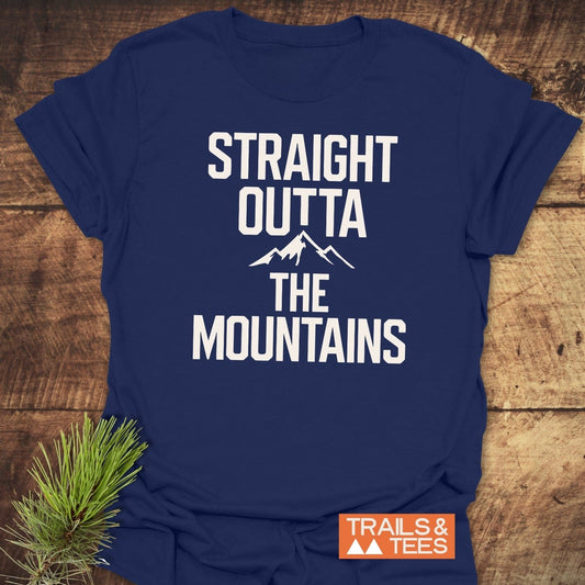 Navy-blue Straight Outta The Mountains T-shirt with white slogan, on wood surface, featuring Trails & Tees logo, pine sprig beside it.