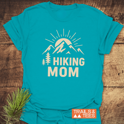 Hiking Mom Sunrise T-Shirt with a mountain and pine graphic on turquoise-blue fabric, laid on wood, showcasing outdoor-themed design with durable, comfortable cotton.