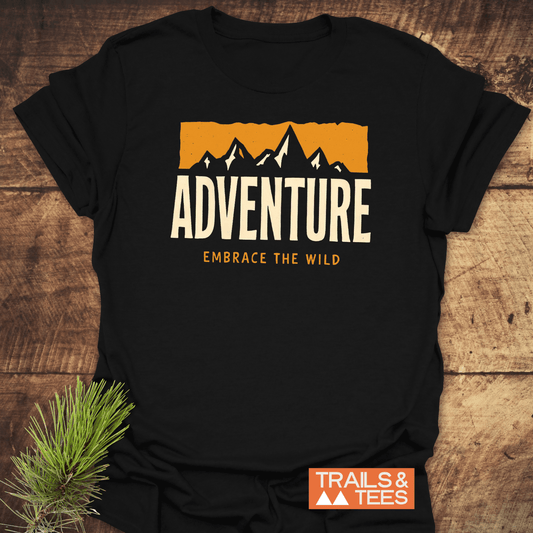 Adventure T-Shirt with mountain graphic and EMBRACE THE WILD tagline, laid on wood. Features orange TRAILS & TEES logo, ideal for outdoor enthusiasts.