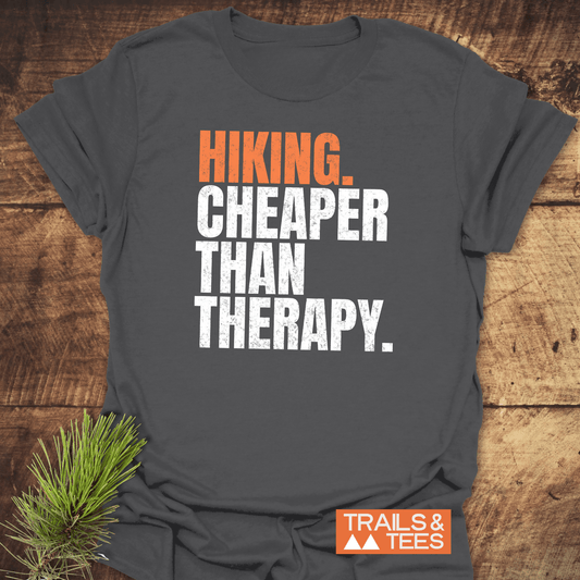 Heather-dark gray T-shirt with HIKING. CHEAPER THAN THERAPY text, orange TRAILS & TEES logo, and pine sprig, highlighting outdoor adventure appeal.