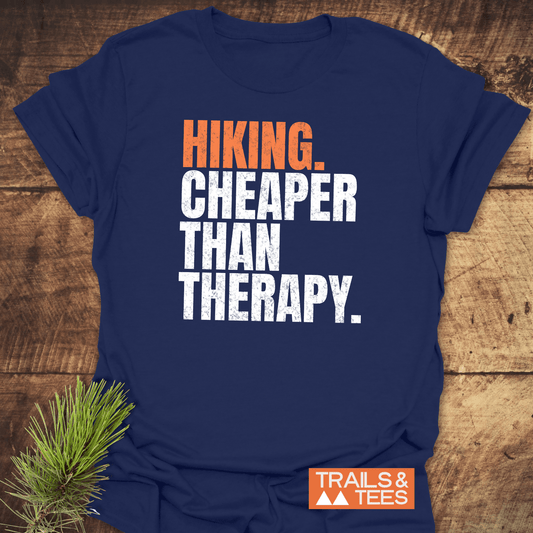 Navy-blue Hiking. Cheaper Than Therapy T-shirt with orange and white lettering, featuring a small TRAILS & TEES logo. Laid on wood with pine sprig.