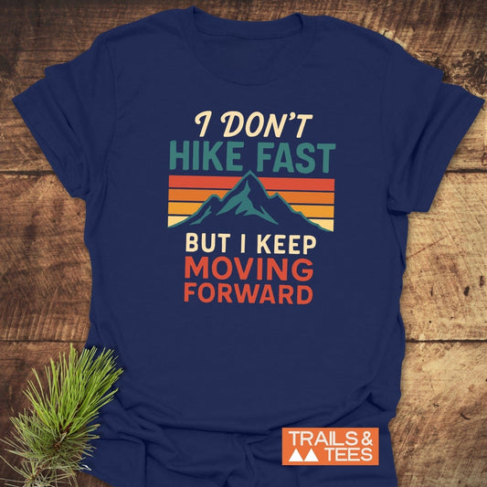 Navy-blue I Don't Hike Fast T-Shirt with mountain and sunset design, on a wooden surface beside a pine sprig; logo TRAILS & TEES visible.