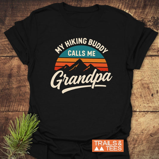Grandpa Hiking Buddy T-Shirt: Black tee with mountain graphic and retro sun, featuring the phrase My Hiking Buddy Calls Me Grandpa; styled with a pine sprig.