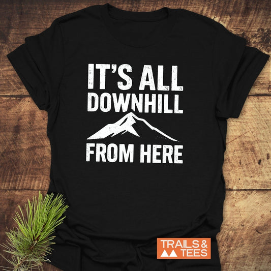 Black All Downhill T-Shirt with mountain graphic and text, laid on wood. Features durable print, ring-spun cotton, and eco-friendly production by Trails & Tees.