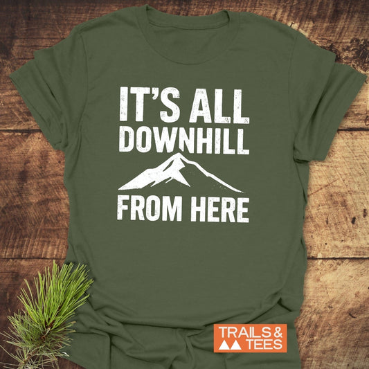 Forest-green All Downhill T-Shirt with distressed white text and mountain design, featuring TRAILS & TEES logo, laid flat on wood, pine sprig nearby.