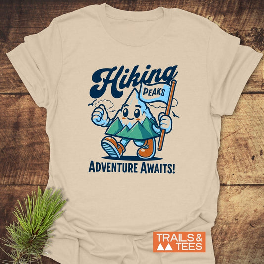 Hiking Time Funny T-Shirt: Cream tee with cartoon mountain character, Hiking script, Adventure Awaits! slogan, TRAILS & TEES logo, laid on wood with pine sprig.