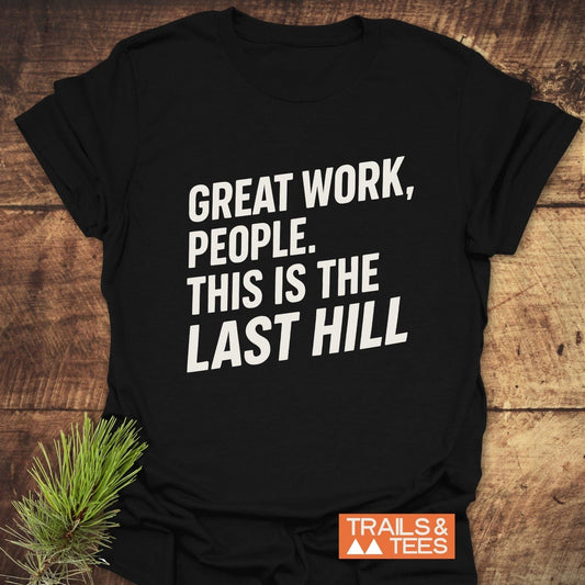 Great Work Funny T-Shirt on wooden surface with slogan, “GREAT WORK, PEOPLE. THIS IS THE LAST HILL,” orange “TRAILS & TEES” logo, pine branch accent.