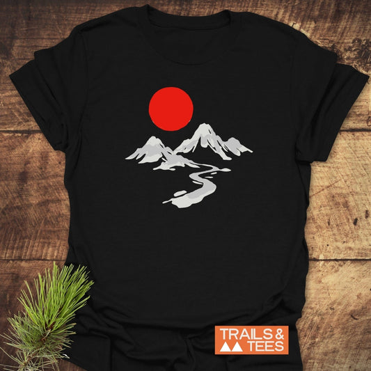Crimson Sun T-Shirt featuring a minimalist outdoor scene with mountains, a river, and a red sun; styled on wood with pine sprig and Trails & Tees logo.