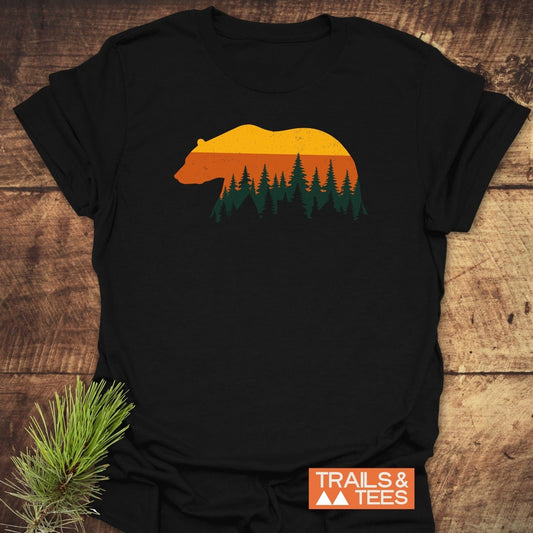 Vintage Grizzly Sunset T-Shirt on wood, featuring a bear silhouette with sunset hues and forest scene. Includes TRAILS & TEES logo and pine branch accent.